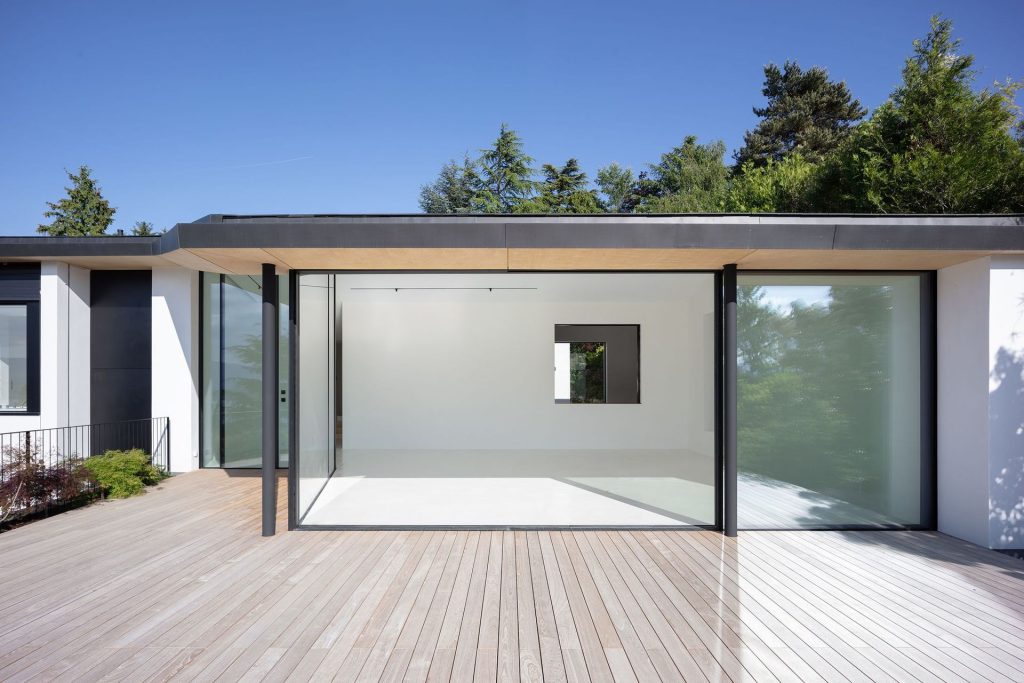 Modern house exterior with sliding glass walls opening to a wooden deck, blending indoor and outdoor living with clean minimalist design.